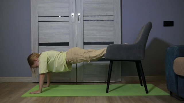 Young adult woman performs challenging decline pushups using stable chair for feet elevation during intense indoor fitness workout session on green yoga mat
