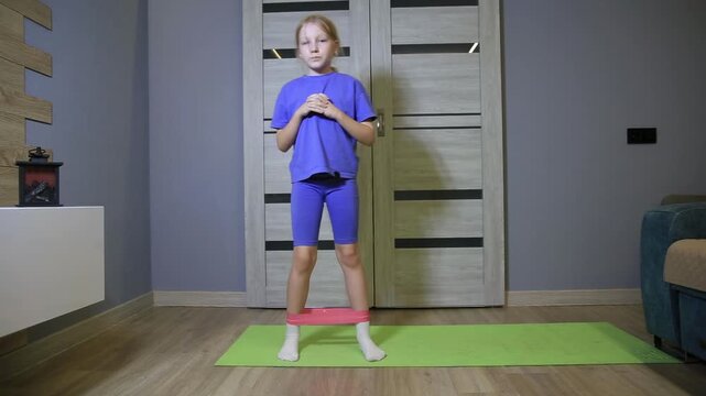 Little girl performing leg exercise with resistance band on yoga mat in living room. Fitness training for child at home, promoting active lifestyle and strength development.