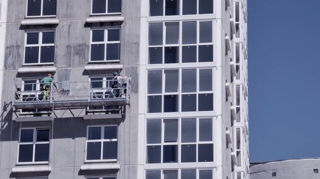 Construction workers perform facade installation and maintenance from suspended platform on unfinished modern high-rise building against clear blue sky.
