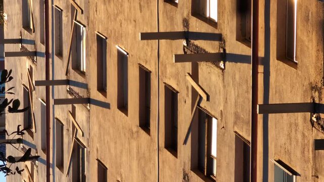 Abstract view of apartment building facade at sunset with geometric shadows from protruding beams and windows, highlighting urban architecture texture and warm light play on aged concrete surface