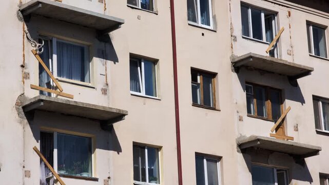 Old residential building facade multiple windows some open some closed damaged concrete balcony slabs visible wooden supports exterior work apartment block structure.