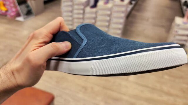 Hand holding blue slip-on canvas shoe with white rubber sole in retail store environment, showcasing footwear for casual wear, comfort, and summer fashion