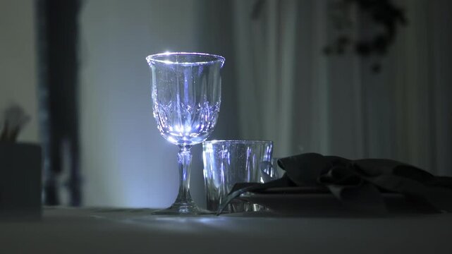 Illuminated crystal wine glass and short tumbler glass on table setting, detail of elegant dinnerware with sparkling light reflection, luxury event atmosphere slow motion