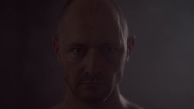 Intense close up portrait of bald man staring directly into camera in low key dark studio. Caucasian male with serious expression and fierce gaze surrounded by dim light and light smoke.