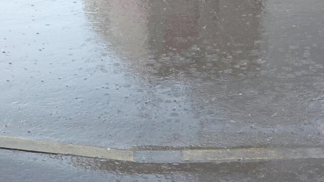 Rain falls heavily on the street in a city, creating puddles and ripples in the water at midday