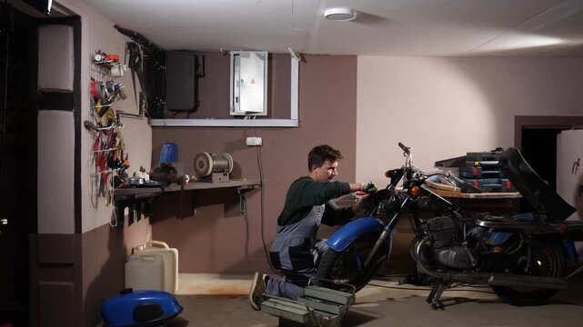 Male mechanic working on a classic motorcycle in a cluttered home garage, using various tools to perform maintenance and diy repair on the engine and frame