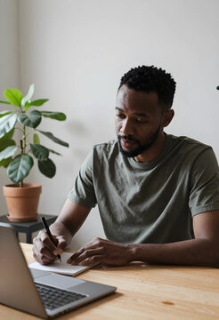 African american man writing notes in a notebook at a wooden desk with a laptop and houseplant, the man is focused on his task, engaged in work or study in a home office environment