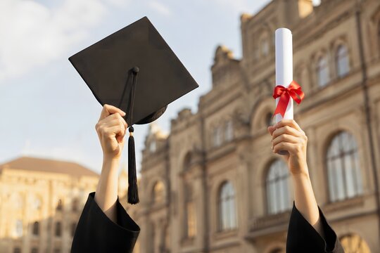 Close up of graduate hands raising cap and diploma against building backdrop