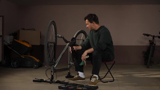 Man holding beer bottle, drinking during a break from working on an upturned bicycle, with a toolbox and various tools open on the concrete floor of a garage