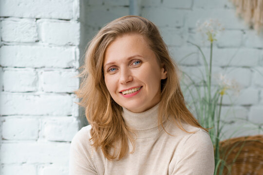 Caucasian woman with light blonde hair smiles with open even white teeth in a beige turtleneck, lit by soft daylight in a bright loft room.