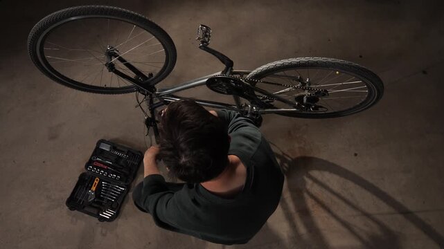 Man from a top view angle taking a break and drinking from a glass bottle while repairing a bicycle in a garage, with a set of tools open on the floor next to him