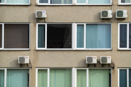 Concrete building facade with office windows, vertical blinds, and air conditioning units mounted on the wall in Talsi, Latvia.