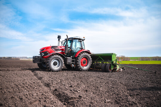Red tractor pulling a modern seeder across a field during the planting season. Realistic agricultural scene showing active fieldwork and modern farming machinery.