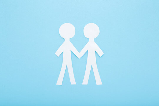 Cut human shapes from white paper on light blue table background. Pastel color. Two people holding hands together. Social relationship concept. Closeup. Flat lay. Top down view.