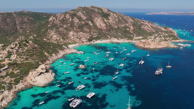 Wide overhead aerial of luxury sailing yachts in turquoise blue water, with a beautiful sandy beach nestled in granite cliffs on the Isola di Spargi in the Maddalena Archipelago in Sardinia, Italy.