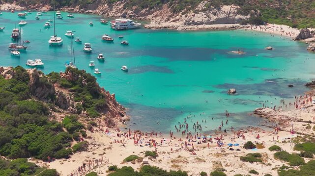 Telephoto aerial of tourists on a beautiful sandy beach nestled in granite cliffs by luxury sailing yachts in turquoise waters of the Isola di Spargi in the Maddalena Archipelago in Sardinia, Italy.