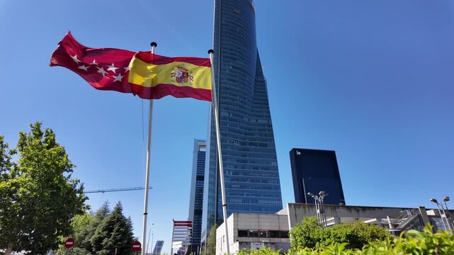 Spanish and Madrid regional flags blowing strongly in the wind with a tall glass office tower in the background.