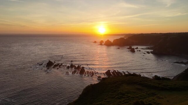 Panoramic drone view of a glowing sunset above a rocky Asturian bay, with layered sea stacks, cliffs and calm coastal water.