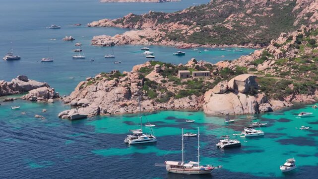 Telephoto aerial of luxury sailing yachts and tour boats, revealing a beautiful sandy beach nestled in granite cliffs on the Isola di Spargi in the Maddalena Archipelago in Sardinia, Italy.