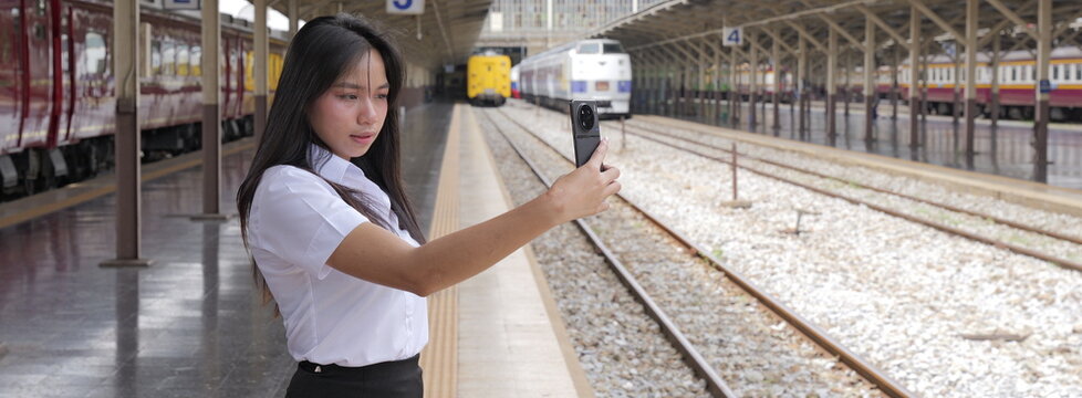A young Asian woman with long black hair in a white short-sleeved university uniform and black skirt takes a selfie with her smartphone