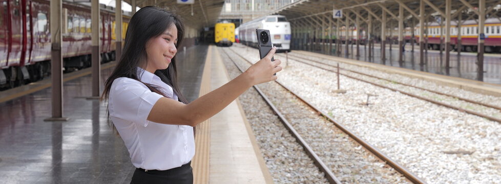 A young Asian woman with long black hair in a white short-sleeved university uniform and black skirt smiles while taking a selfie with her smartphone