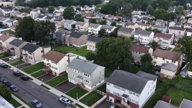 Grid of single-family houses fills dense residential neighborhood in New Jersey, tree-lined streets and parked cars line rows of white and gray rooftops throughout. Aerial drone flyover shot