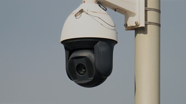 Close-up motion of a modern outdoor security camera rotating on a pole against a clear sky.