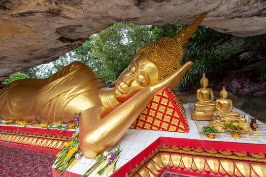 Side view of a large golden reclining Buddha statue resting beneath a giant rock cliff alongside smaller Buddha statues and offerings.