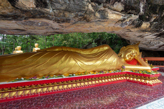 Golden reclining Buddha statue situated under a natural rock cliff overhang in a Thai temple, adorned with fresh flower offerings.