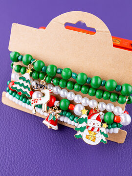 Close-up of a set of green and white Christmas bracelets with pendants in the shape of a snowman, reindeer, and Christmas tree on purple background