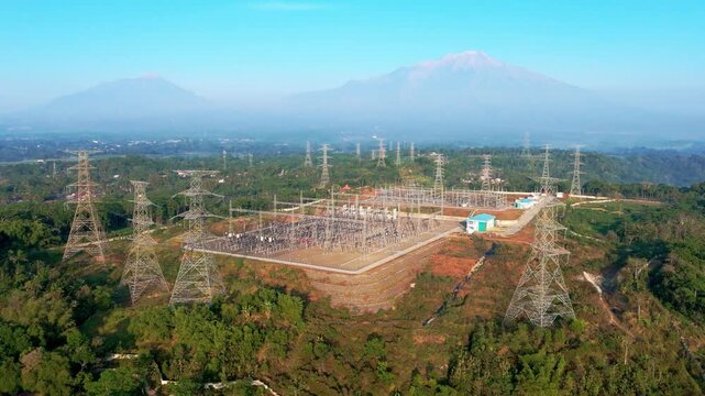 Aerial view of Ampel 150kV Substation with Mount Merbabu and Mount Merapi in the background, surrounded by lush green hills in Kecamatan Ampel, Jawa Tengah, Indonesia.