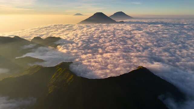Aerial view of Mount Bismo peak rising above a vast sea of clouds during golden hour with soft sunlight illuminating the mountain slopes in Kecamatan Kejajar, Jawa Tengah, Indonesia.