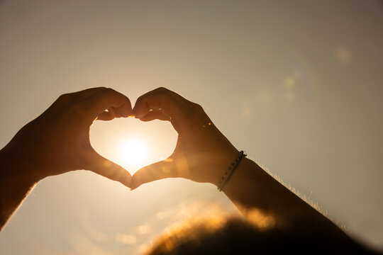 Silhouette of hands forming a heart shape around the bright sun against sky.