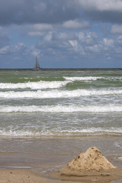 Mound of sand on a sandy beach with foamy waves and a distant sailboat under cloudy skies at Cadzand - The Netherlands