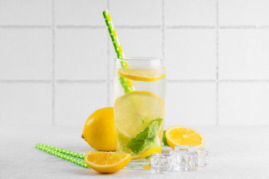 Refreshing homemade lemonade served in clear glass with fresh lemon slices, mint leaves, ice cubes and paper straws on a light kitchen counter against a white tile background