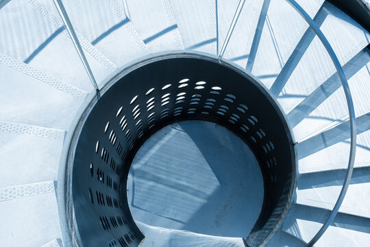 Viewed downward, a spiral staircase opens through the interior around a central void, where blue circular geometry surrounds steps beside a white railing, creating symmetry