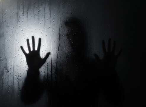 A silhouette of a person standing behind a rainy glass window in the dark