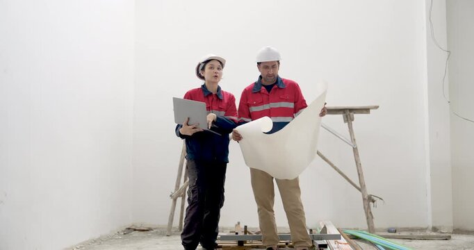 Asian woman engineer and male contractor review blueprint beside survey tripod, discussing construction site safety inspection, building project progress, document checking, quality control indoors.