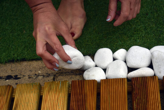 Close-up of a woman's hands arranging white decorative pebbles along a garden border beside a wooden deck and artificial grass. Outdoor landscaping and garden decoration concept.