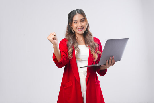 Excited young Asian woman raising her fists with happy successful expression while holding laptop.