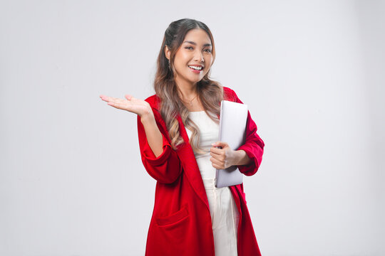 Cheerful young Asian woman in red jacket holding laptop while presenting copy space behind her.