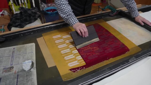 Craftsman drawing a squeegee across an ochre silkscreen, forcing crimson pigment through the stencil onto the sheet below