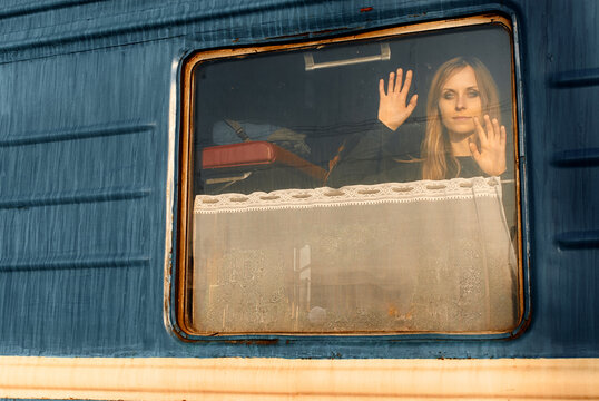A young woman presses her hands to a train window and gazes outward with a reflective, melancholic expression as luggage sits behind her, evoking travel, farewell and quiet solitude.