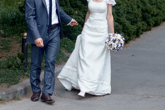 Bride and groom walk together outdoors along a paved path; the bride holds a white lace wedding dress and a purple-white bouquet, evoking romance, celebration, partnership and intimacy.