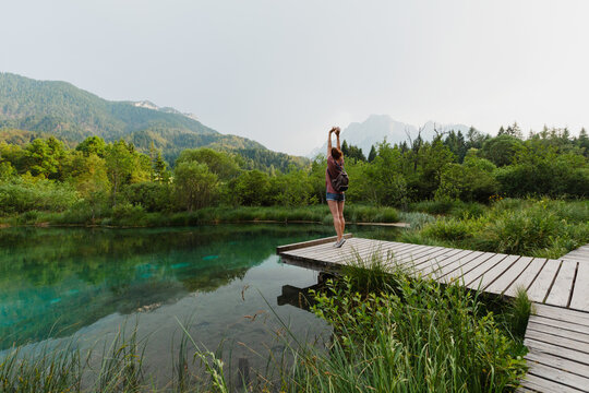 A female traveler arms outstretched while standing on a wooden path beside an emerald lake. Vitality and eco-tourism concept showcasing physical well being immersed in a pristine environment.