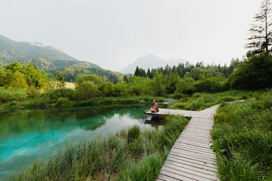 A woman practices outdoor meditation on a wooden boardwalk beside an emerald alpine lake. Harmonious eco-tourism concept focusing on spiritual connection with beautiful natural landscapes.