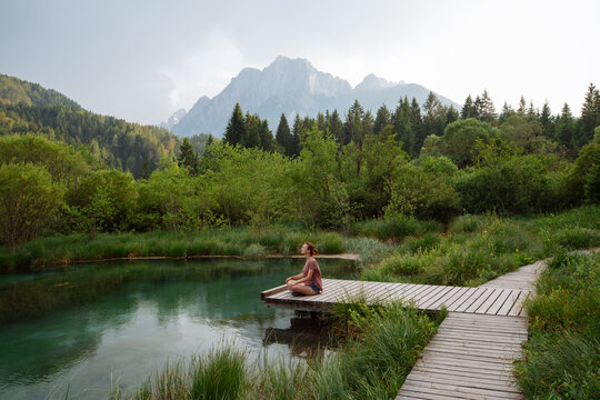A woman practices outdoor meditation on a wooden boardwalk beside an emerald alpine lake. Harmonious eco-tourism concept focusing on spiritual connection with beautiful natural landscapes.
