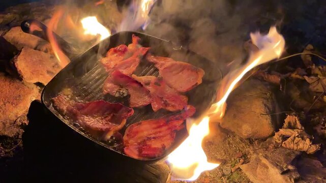 Static shot of bacon cooking in cast iron pan over open campfire in the dark at night.