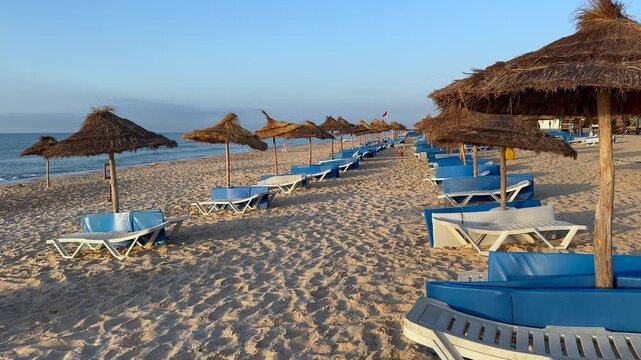 Rows of blue sun loungers and straw umbrellas line an empty sandy beach in soft early morning light on Hammamet beach, Tunisia.