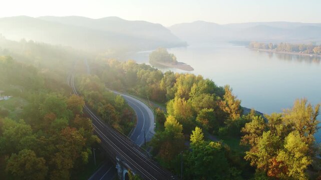 Wide aerial shot flying over a stone railway bridge and curved coastal road alongside the calm, misty Danube river, surrounded by autumn forests at sunrise.